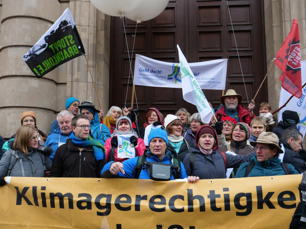 Eine Gruppe von Menschen mit einem Banner, Fahnen und Luftballons vor einem Gebäude mit Säulen und einer Tür während eines Klimastreiks in Deutschland.