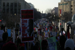Menschen protestieren auf einer Straße und halten Schilder, im Hintergrund sind Gebäude, Bäume, Laternenpfähle und der Himmel zu sehen.