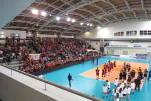 Eine Gruppe von Menschen spielt Volleyball in einer Turnhalle mit einem Netz, umgeben von einer Absperrung, Zuschauern auf Stühlen mit Schildern und einer Wand mit Fenstern und Deckenleuchten im Hintergrund.