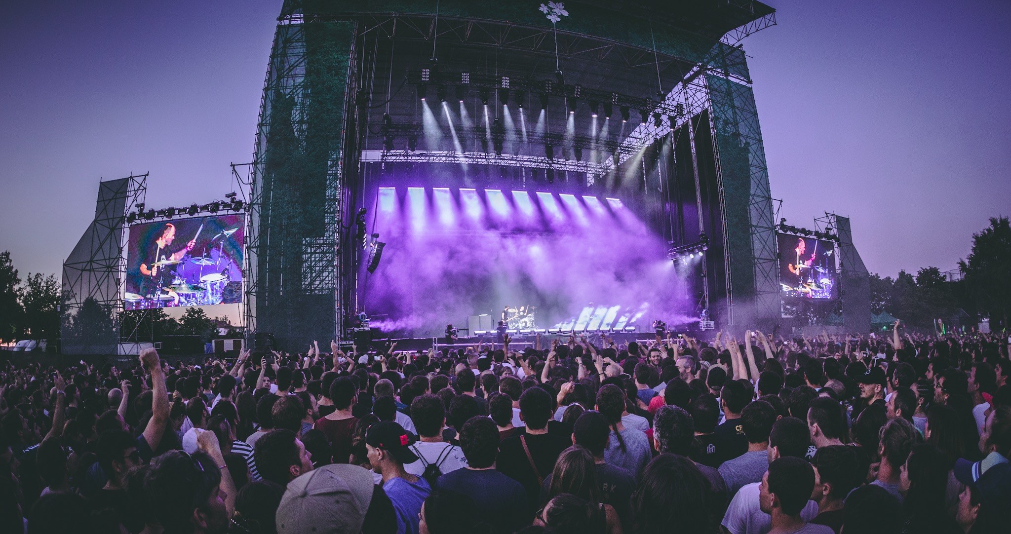 Eine große Menschenmenge auf einem Musikfestival mit einer Bühne, auf der Musiker Instrumente spielen, beleuchtet von Scheinwerfern und umgeben von Bildschirmen, vor einer Kulisse aus einem Gebäude, Bäumen und einem klaren blauen Himmel.