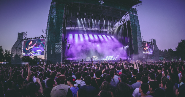 Eine große Menschenmenge auf einem Musikfestival mit einer Bühne, auf der Musiker Instrumente spielen, beleuchtet von Scheinwerfern und umgeben von Bildschirmen, vor einer Kulisse aus einem Gebäude, Bäumen und einem klaren blauen Himmel.