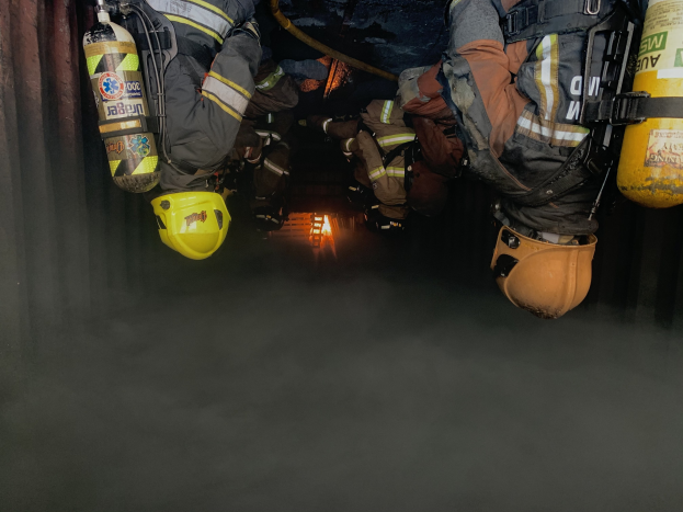 Feuerwehrleute in Helmen und Jacken mit Atemschutz arbeiten in einem raucherfüllten Tunnel, mit einer Lichtquelle im Hintergrund.