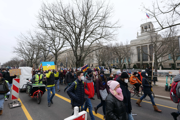 Eine große Gruppe von Menschen marschiert bei einer Demonstration auf einer Straße in Washington, D.C., mit Schildern und Bannern, einige fahren Fahrräder, unter einem klaren blauen Himmel.