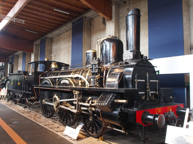 Alte Dampflokomotive in einem Museums Setting mit Eisenbahnschienen, Informationsschildern und anderen Ausstellungsstücken im Hintergrund.