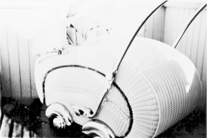 Ein Schwarz-Weiß-Foto eines Babys in einem Kinderwagen, das eine weiße Strampelanzug trägt und friedlich schaut, mit einer Wand im Hintergrund.