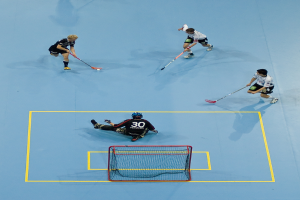 Drei Personen in Sportkleidung spielen Hockey, wobei eine Person auf der rechten Seite den Ball mit einem Hockeystock schlägt und eine weitere Person hinter einem Netz auf dem Boden sitzt.