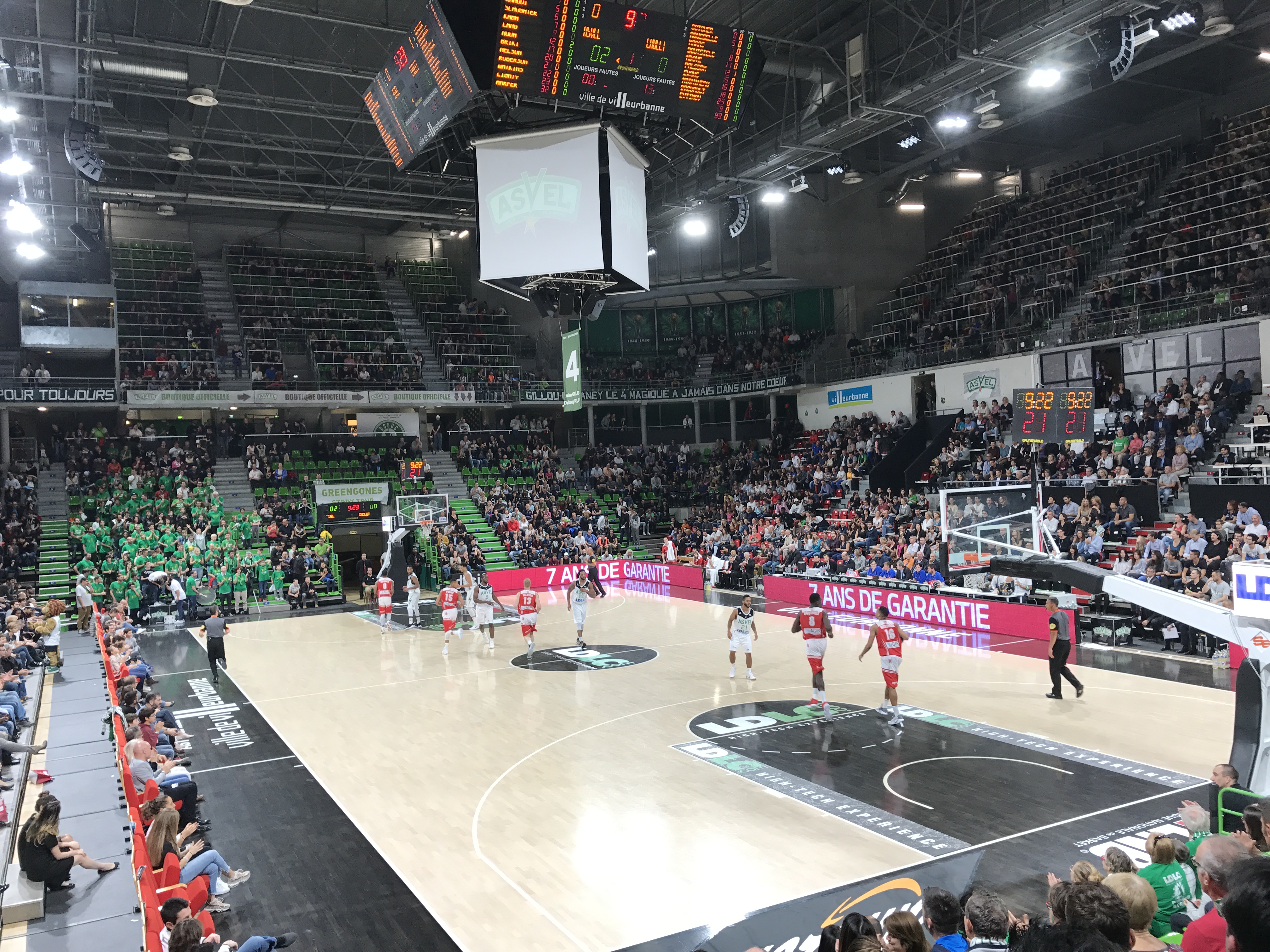 Ein Basketballspiel in einer großen Arena mit Zuschauern in den Rängen, Anzeigetafeln mit "Asvel vs Galatasaray" und einer Decke mit Lampen und einem Bildschirm.