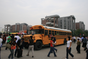 Eine Gruppe von Menschen, einige mit Taschen, geht vor einem Schulbus auf einer Straße mit Gebäuden, Bäumen und Laternen unter einem klaren blauen Himmel.