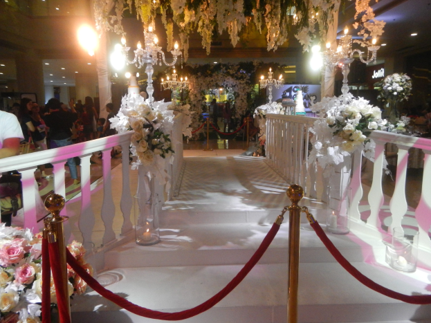 Eine Trauung in einem Einkaufszentrum mit weißem Teppich, verzierten Geländern, Blumenarrangements und Deckenbeleuchtung, bei der eine Menge Menschen mit Taschen anwesend sind.