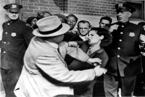 Zwei Personen kämpfen in einem Schwarz-Weiß-Bild, mit mehreren anderen im Hintergrund, einige tragen Mützen und eine Wand im Hintergrund.