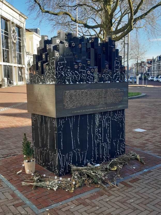 Denkmal in einem Stadtplatz gewidmet an Holocaust-Opfern, mit einem Baum davor, einem Topfgewächshaus auf dem Boden, verstreuten trockenen Blättern und Gebäuden, Fahrzeugen, Masten und Bäumen im Hintergrund.
