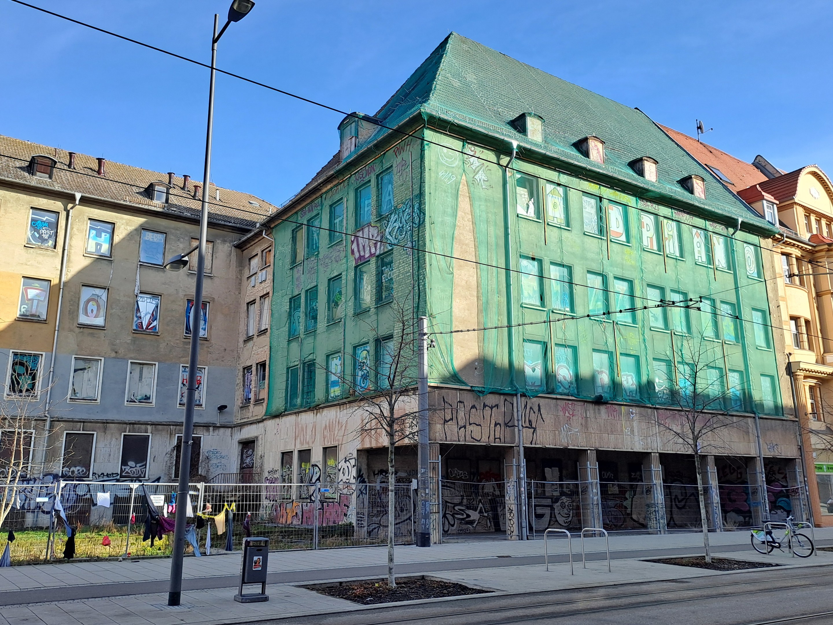 Große grünes Gebäude mit Graffiti an seiner Seite, Teil des Berlin Wall Project in Berlin, Deutschland, umgeben von urbanen Elementen wie Straßenlaternen, Fahrzeugen, Passanten und Bäumen unter einem bewölktem Himmel.