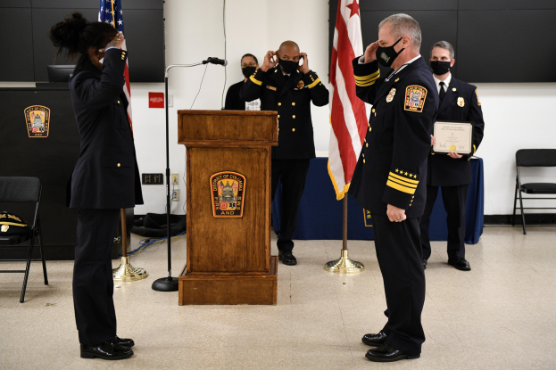 Gruppe uniformierter Personen in Masken vor einem Rednerpult bei einer Vereidigung, eine hält ein Zertifikat, umgeben von Flaggen und Stühlen.