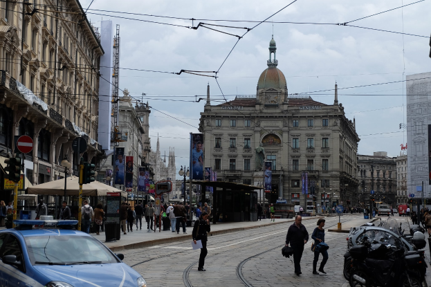 Eine belebte Stadtstraße mit einem geparkten Polizeiauto, Fußgängern mit Taschen, fahrenden Fahrzeugen, Gebäuden mit Fenstern und Bannern, Laternen, Verkehrsampeln und einem bewölkten Himmel.