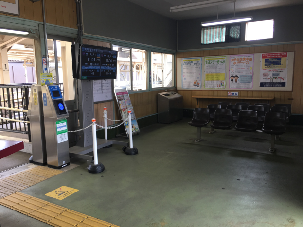 Ein belebter Bahnhofseingang mit Sitzgelegenheiten, einer Informations counter, Ticketautomaten, Kettenzäunen, Informationsanzeigen, Wandplakaten, Deckenbeleuchtung und Geländern.