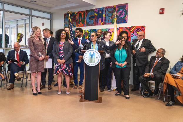 Gruppe von Menschen in einem Raum mit einem Podium, Mikrofon und einer Tafel mit der Aufschrift "Sancho-Stadtrat tritt dem Stadtrat bei", umgeben von Fahnen, Sitzplätzen und einem Feuerlöscher.