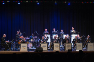 Eine Gruppe von Musikern auf der Bühne, die Instrumente spielen, mit Mikrofonen und Ständern, Schildern mit der Aufschrift "The New Orleans Jazz Band", ein Vorhang im Hintergrund und ein Publikum darunter unter Bühnenlichtern.