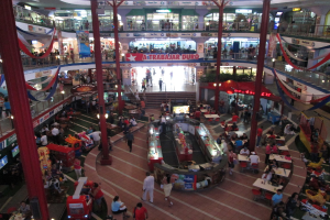 Eine belebte Einkaufszentrumszene mit Säulen, Menschen, Tischen, Fliesenboden, Flaggen, Geschäften, einem Banner, Treppen und einem Namensschild.