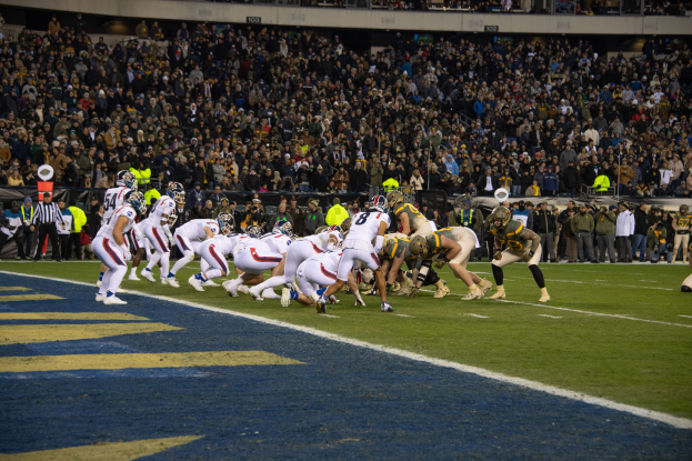 Ein Fussballspiel im Gange mit Spielern in Sportbekleidung und Helmen auf dem Feld, Zuschauern, Gel"andern und Tafeln mit Text im Hintergrund.