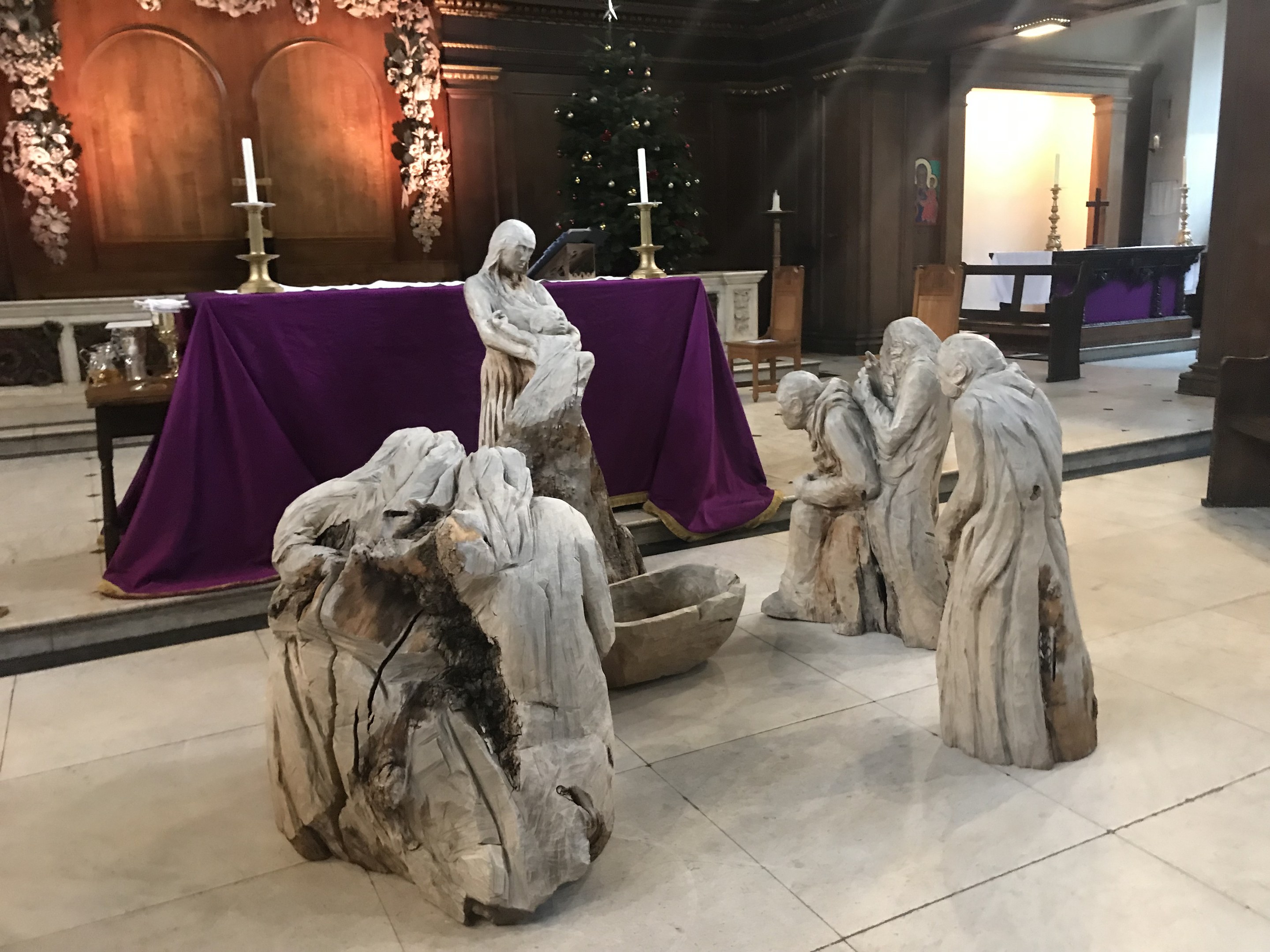 Krippe in einer Kirche mit Statuen auf dem Boden, einem Tisch mit einer Decke und Kerzenständern, Stühlen, einem geschmückten Weihnachtsbaum mit Lichtern und einer Wand im Hintergrund.