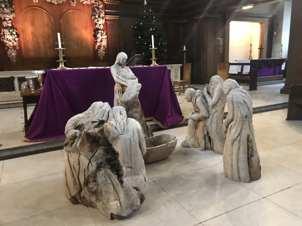 Krippe in einer Kirche mit Statuen auf dem Boden, einem Tisch mit einer Decke und Kerzenständern, Stühlen, einem geschmückten Weihnachtsbaum mit Lichtern und einer Wand im Hintergrund.