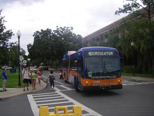 Ein Shuttlebus fährt auf einer Straße mit hohen Gebäuden, Fußgängern auf dem Gehweg, Laternen und Bäumen unter einem bewölkten Himmel.