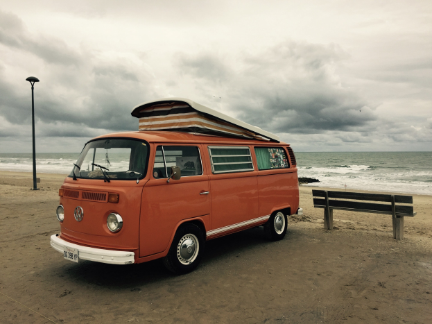 Ein oranger VW-Bus auf einem Strand neben dem Meer geparkt, mit einer Bank und einem Laternenmast in der N├Ąhe, unter einem bew├Âlktem Himmel mit dem Wasser im Hintergrund.