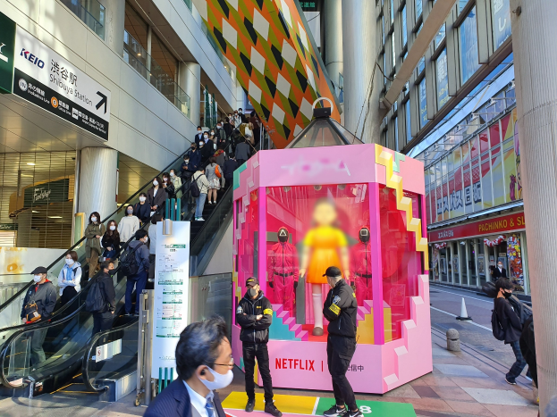 Eine Gruppe von Menschen steht vor einem Gebäude mit Glaswänden, Texttafeln, einige tragen Masken, in der Nähe einer Rolltreppe; ein pinker Kasten in der Mitte zeigt Schaufensterpuppen und trägt "Netflix's neuen Netflix-Stand in Tokyo."