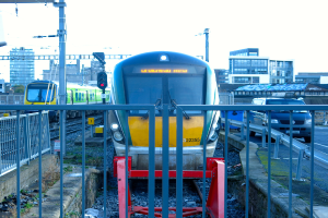 Zwei Züge auf dem Schienenstrang mit einem Zaun im Vordergrund, Masten auf dem Land, einer mit einer Signalleuchte, ein Auto auf der Straße rechts, Gebäude im Hintergrund und der Himmel oben.