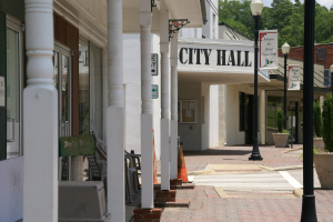 Ein Rathaus umgeben von Geb├Ąuden, Stra├čenlaternen, Stra├čenlaternen, Namensschildern, Kraftfahrzeugen, Verkehrskegeln, St├╝hlen, Topfpflanzen, B├Ąumen und einer bew├Âlktem Himmel.