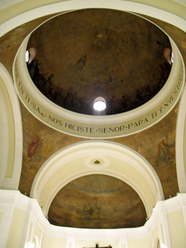 Das Innere einer Kirche mit einem Kreuz an der Decke, beleuchtet von Lichtern und umgeben von Säulen, mit Wänden, die mit Gemälden und Texten geschmückt sind.