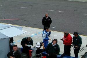 Eine Gruppe von Menschen, die stehen, einige tragen Headsets und schreiben in Büchern, mit einer Straße im Hintergrund und einem kleinen Raum links.