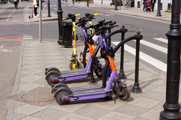 Eine Reihe von E-Scootern, die entlang einer Straße mit Fußgängern, Radfahrern, einem Mülleimer im Vordergrund und Gebäuden, Bäumen und Pflanzen im Hintergrund geparkt sind.