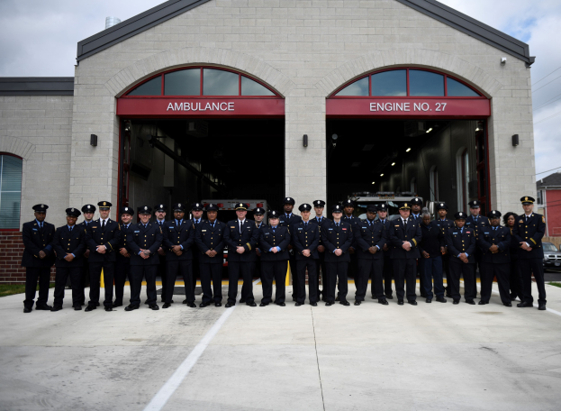 Feuerwehrleute in Uniform und Mützen vor einer Feuerwache mit Fahrzeugen, Gebäuden und einem klaren Himmel im Hintergrund.
