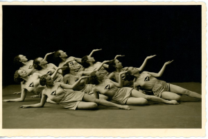 Ein Schwarz-Weiß-Foto einer Gruppe von Frauen in einheitlicher Kleidung, die Boden-Gymnastik machen, mit verschiedenen Arm- und Beinposen, zurückgekämmtem Haar, strahlend und in die Kamera schauend, mit einem unscharfen, verträumten Hintergrund.