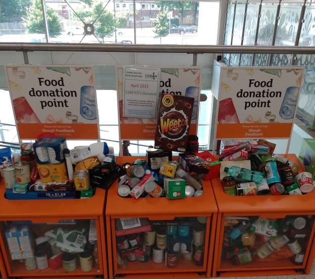 Orange Schränke mit Lebensmittelspenden gefüllt, darunter Flaschen, Dosen und Kartons, mit informativen Schildern im Hintergrund; Glasfenster zeigen Fahrzeuge, Bäume, Gebäude und Himmel.