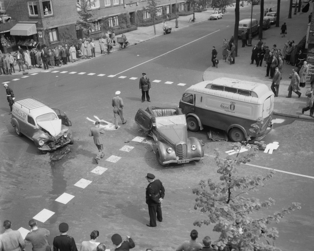 Schwarz-weißes Bild eines Autounfalls an einer Straßenkreuzung mit mehreren Fahrzeugen, darunter ein Lieferwagen, und Passanten; Gebäude, Bäume, Pfosten und Texttafeln sind im Hintergrund zu sehen.