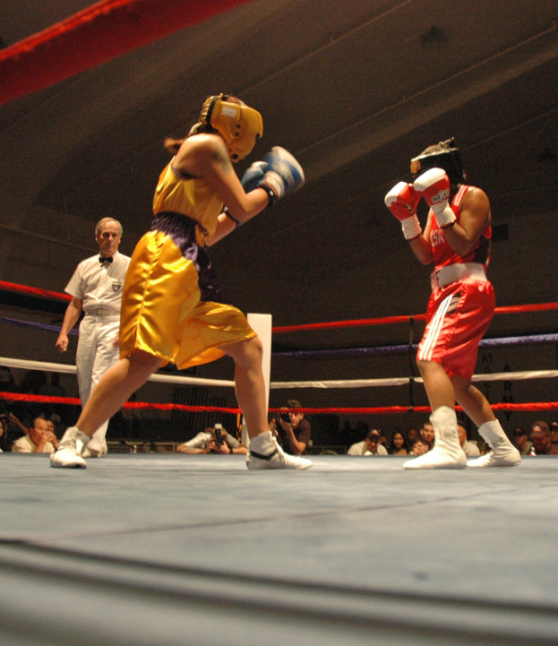 Zwei Boxer kämpfen in einem Ring mit einem Schiedsrichter und Zuschauern, einige mit Kameras, sichtbare Decke darüber.