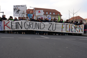 Protestierende halten ein Transparent mit der Aufschrift 'Kein Grund zu Feiern' gegen deutsche Sparmaßnahmen, mit Straßeninfrastruktur und Gebäuden im Hintergrund.