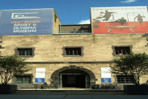 Außenansicht des Deutschen Sport & Olympia Museums in Berlin, das ein Gebäude mit Fenstern und einer Tür zeigt, umgeben von Bäumen und Bannern, unter einem bewölkten Himmel.