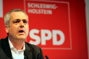 Eine Person in einem schwarzen Mantel steht links im Bild und spricht, mit einem Banner im Hintergrund.