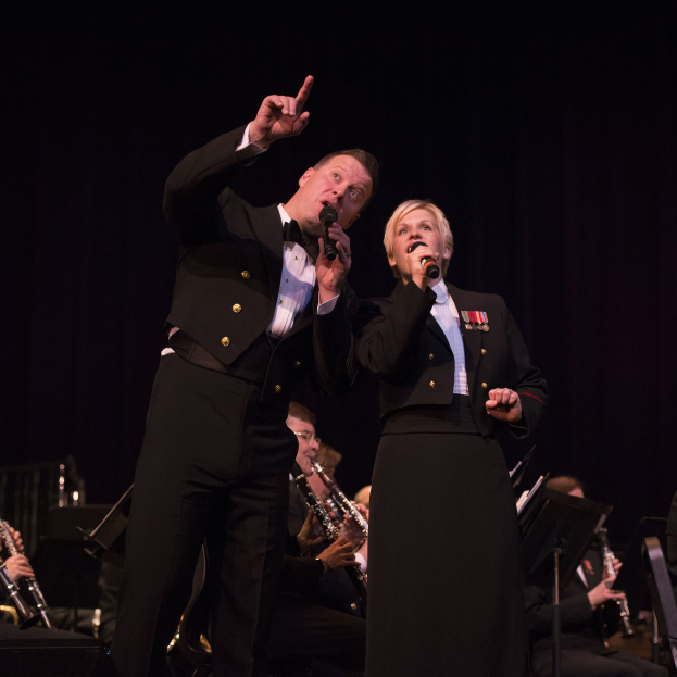 Ein Mann und eine Frau in Fracks stehen auf einer Bühne und halten Mikrofone, mit einem dunklen Hintergrund und einem sitzenden Orchester dahinter.