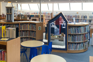 Ein Bibliotheksinneres mit Bücherregalen, Tischen, Stühlen, Sofas mit Kissen, einer Säule und Deckenleuchten.