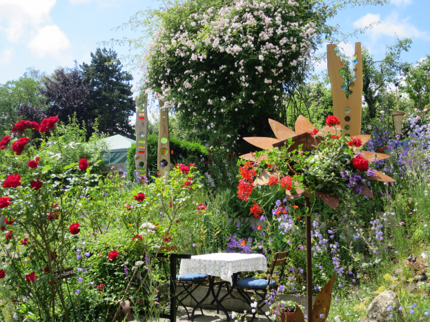 Ein Garten mit bunten Blumen und Pflanzen, der einen zentralen Tisch mit einer Decke und Stühlen zeigt, umgeben von Bäumen unter einem bewölkten Himmel, auf der Chelsea Flower Show.