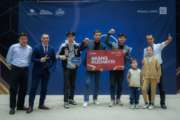 Gruppe von Menschen auf einer Bühne mit einer Tafel, auf der "Akang Kuchaydi - Zakovat Quiz" steht, im Hintergrund ein Banner und ein Strichcode.