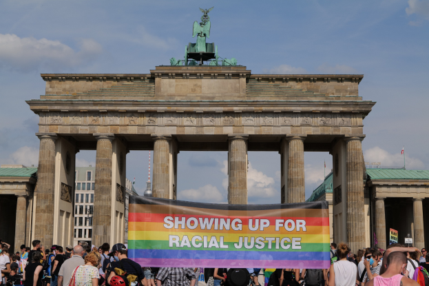 Eine Gruppe von Menschen hält eine "Racial Justice"-Schrifttafel vor dem Brandenburger Tor in Berlin, Deutschland, mit den Säulen und der Statue des Tors im Vordergrund und Gebäuden und einem bewölkten Himmel im Hintergrund.