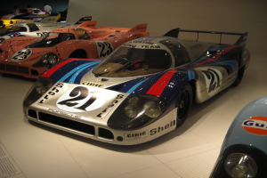Ein Porsche 917K Le Mans Rennauto wird in einem Museum ausgestellt, umgeben von anderen Sportwagen und Menschen, mit einer Wand im Hintergrund und einem Blatt Papier mit Text auf der linken Seite.