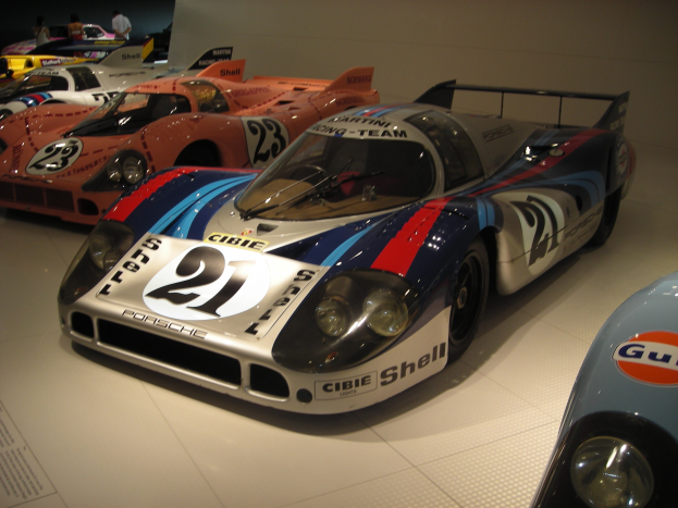 Ein Porsche 917K Le Mans Rennauto wird in einem Museum ausgestellt, umgeben von anderen Sportwagen und Menschen, mit einer Wand im Hintergrund und einem Blatt Papier mit Text auf der linken Seite.