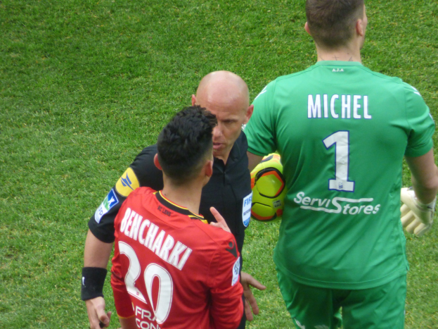 Schiedsrichter spricht mit zwei Fußballspielern auf einem grasbedeckten Feld, ein Spieler hält einen Ball.