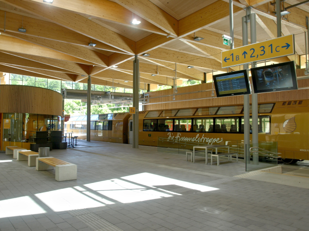 Ein belebter Bahnhof mit einem stehenden Zug auf den Schienen, umgeben von Bänken, Tischen, Pfosten, Schildern, Lampen und Bäumen, während Menschen auf den Zug warten.